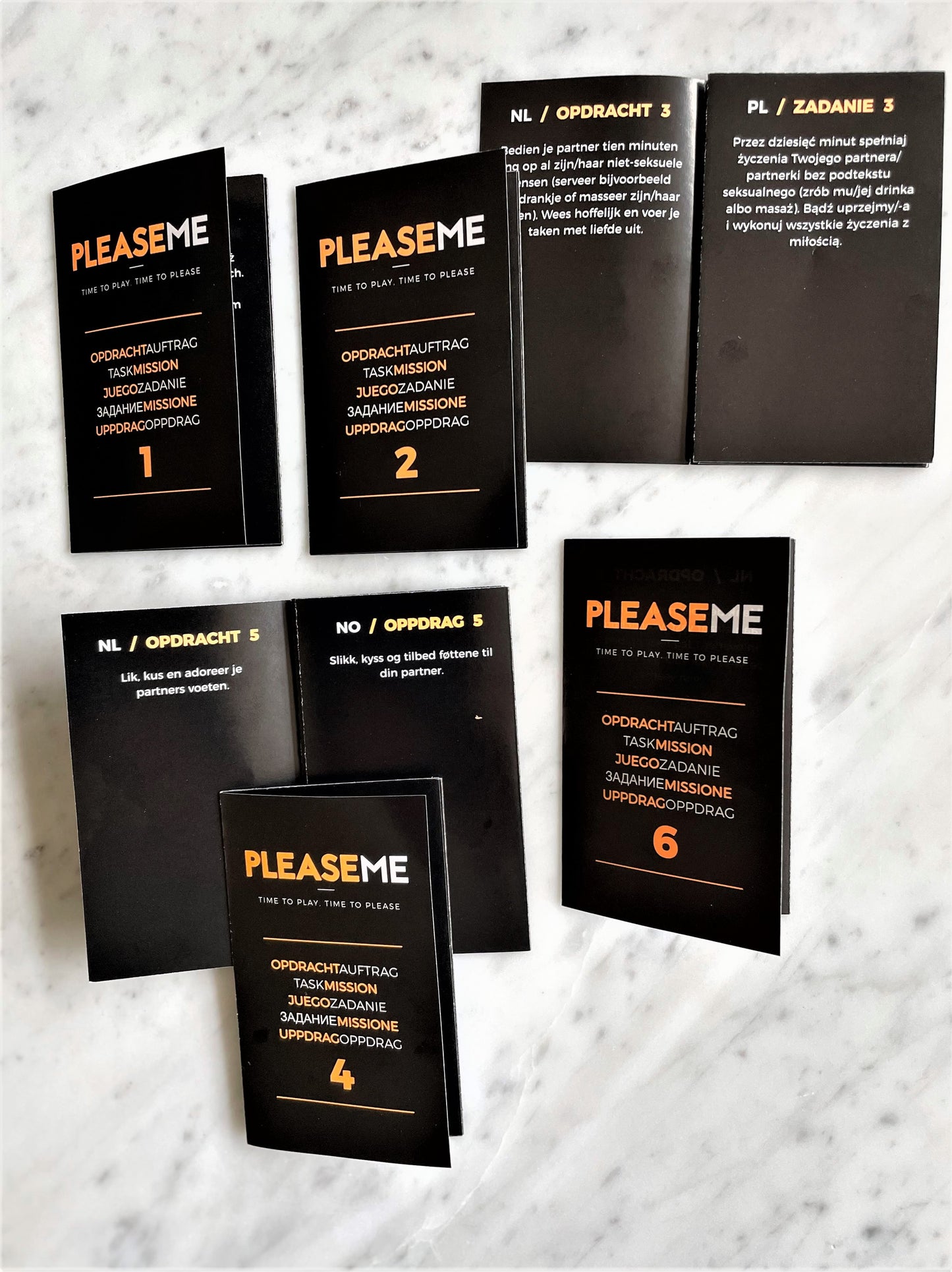 PleaseMe opdrachtkaarten uitgespreid op marmer, genummerd 1-6, zwart met oranje tekst