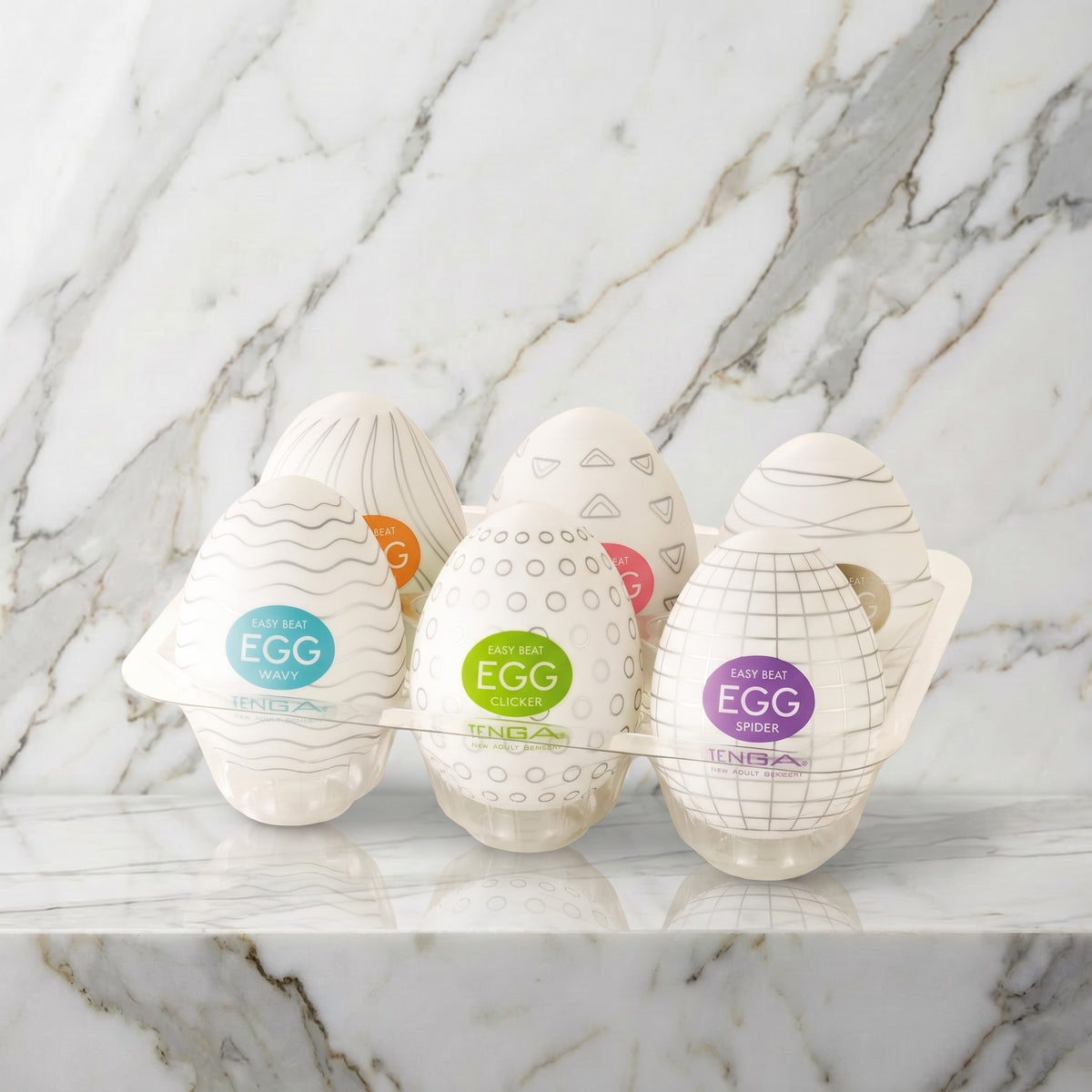 Zes witte Tenga Eggs met gekleurde labels in een doorzichtig houdertje op een marmeren plank