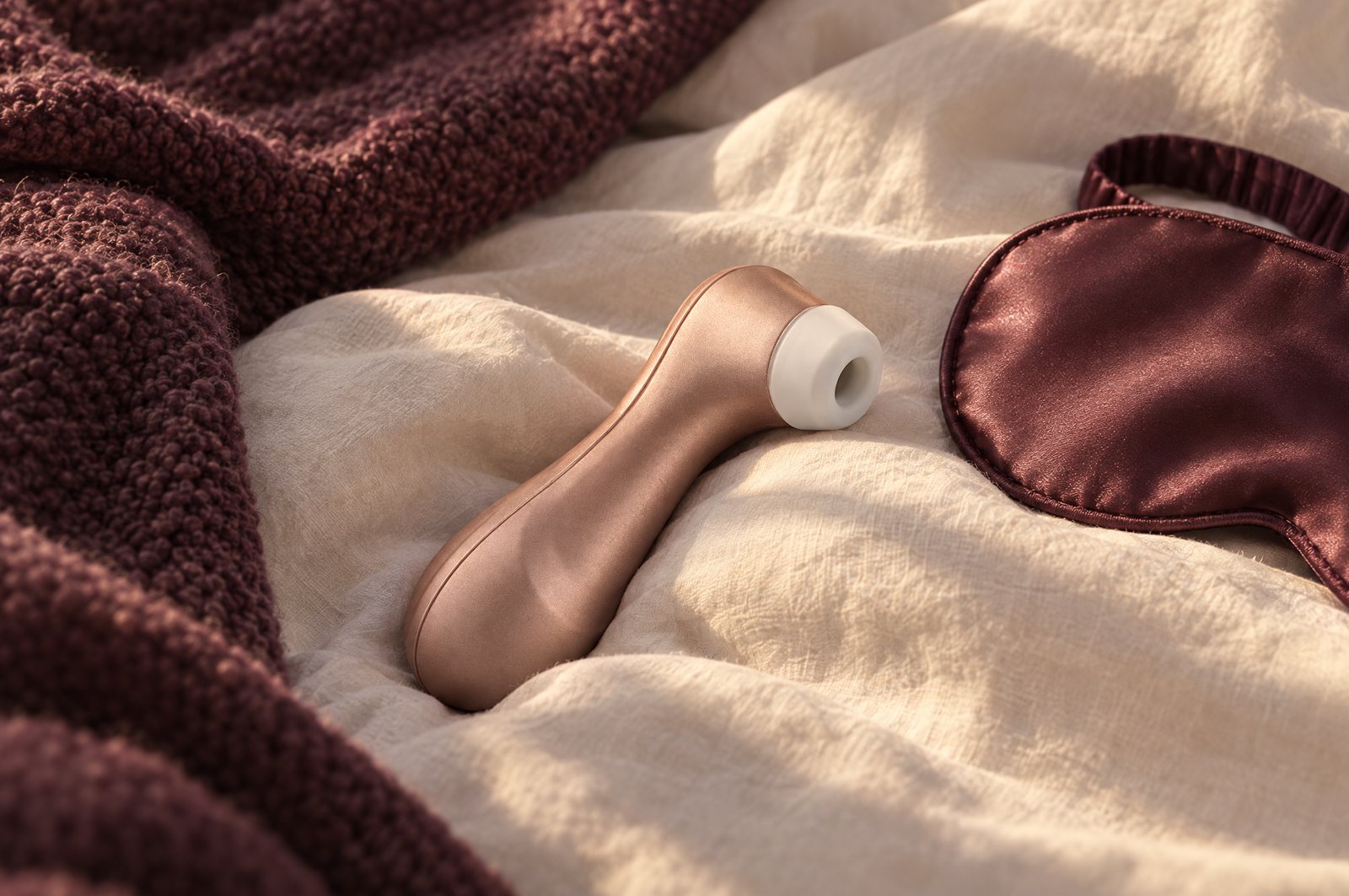 Satisfyer Pro 2 ligt op een opgemaakt bed naast een gebreide plaid en een satijnen oogmasker