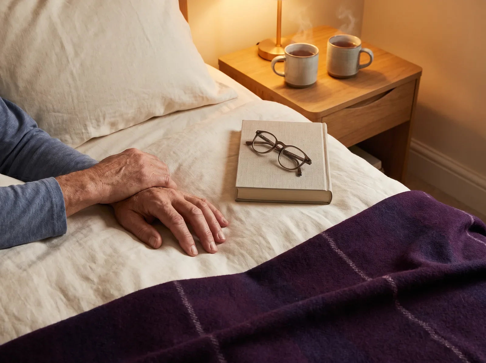 Sfeerbeeld: twee handen naast een boek met leesbril op bed, met thee bij lamplicht