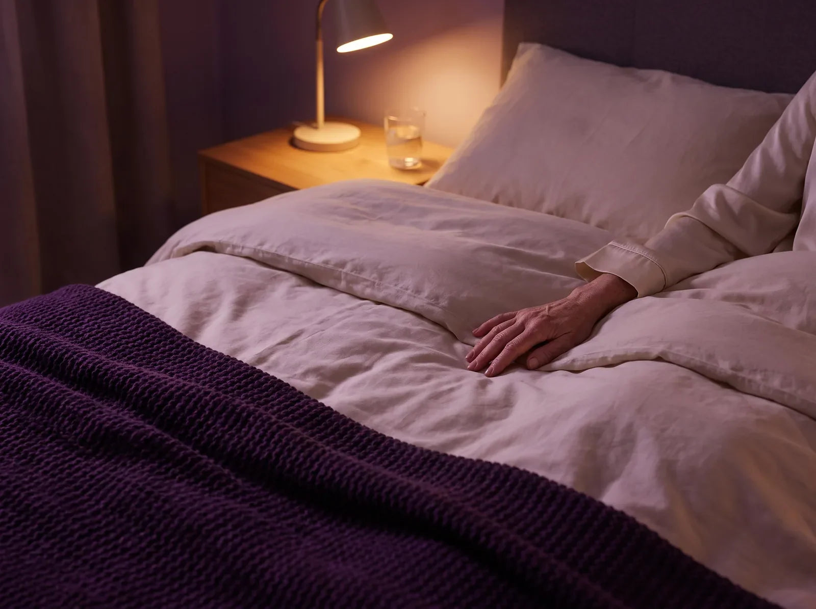 Sfeerbeeld: een hand rust 's nachts op het dekbed, bij het licht van een leeslamp