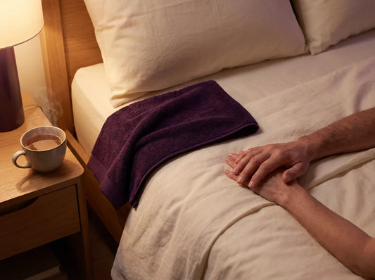 Sfeerbeeld: twee handen op een opgemaakt bed naast een donkerpaarse handdoek, bij warm lamplicht
