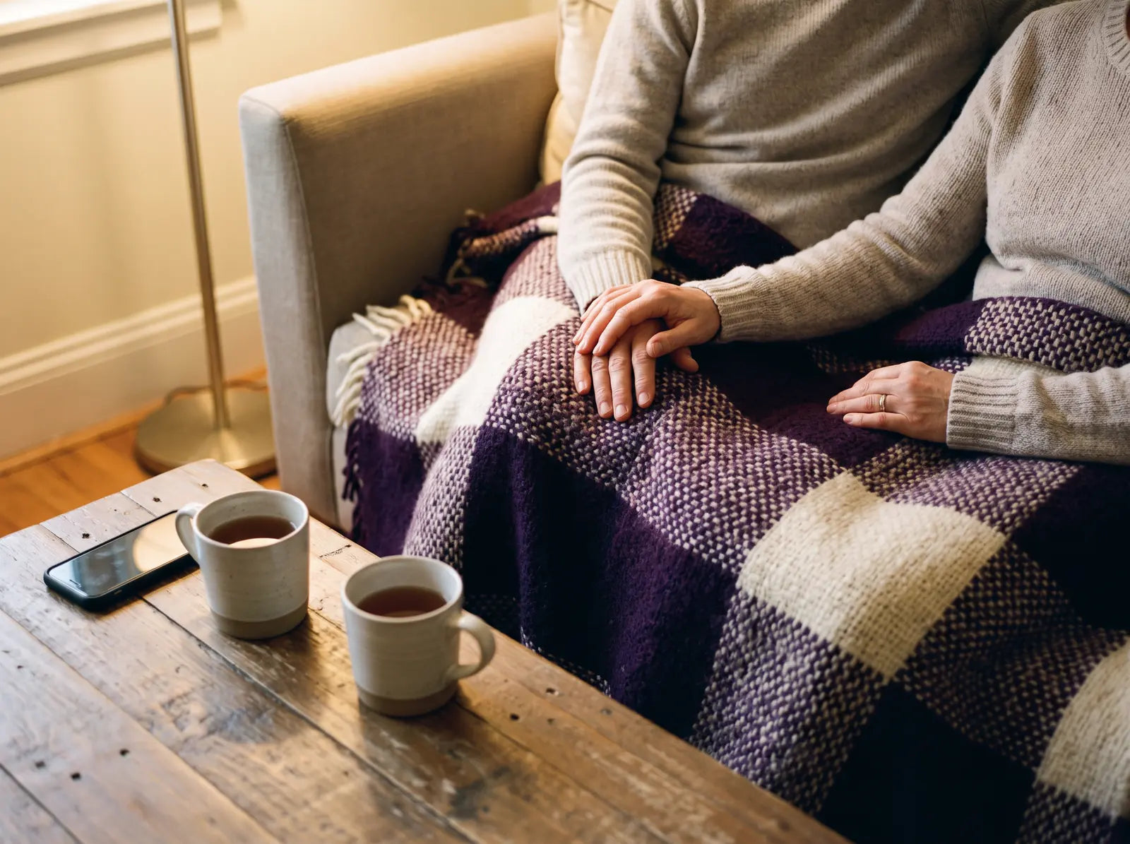 Sfeerbeeld: twee handen rusten in elkaar op een paarse plaid, met thee op tafel