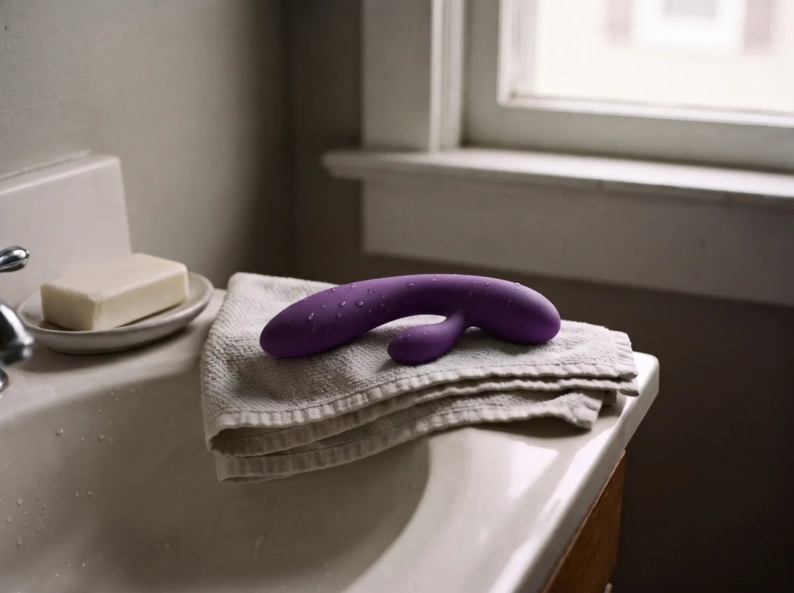 Sfeerbeeld: een net afgespoelde paarse rabbitvibrator droogt op een opgevouwen handdoek naast de wastafel