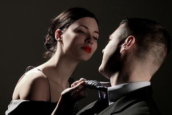 Een elegante vrouw met rode lippen buigt zich verleidelijk naar een man in een pak, terwijl ze zijn stropdas vastpakt in een intiem, spanningsvol moment tegen een donkere achtergrond.