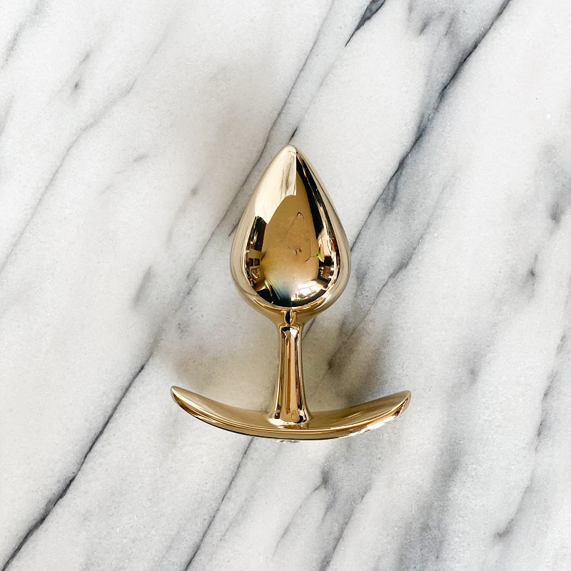 Goudkleurige metalen buttplug met brede gebogen basis, van boven gezien op een marmeren plank
