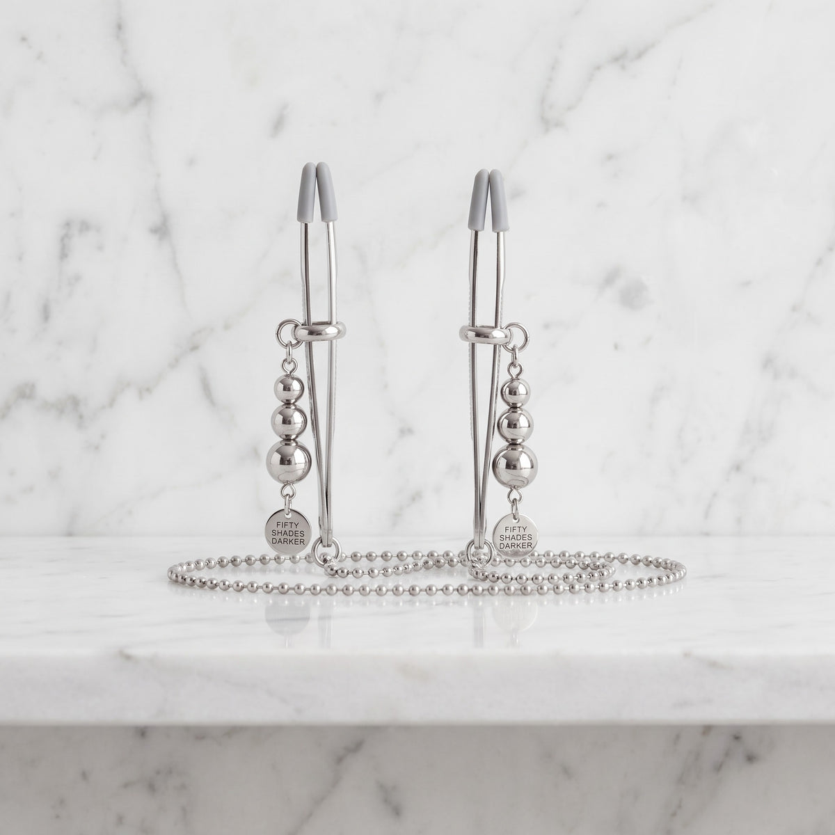 Twee zilveren tepelklemmen met bolletjes en kogelketting op een marmeren plank, Fifty Shades Darker