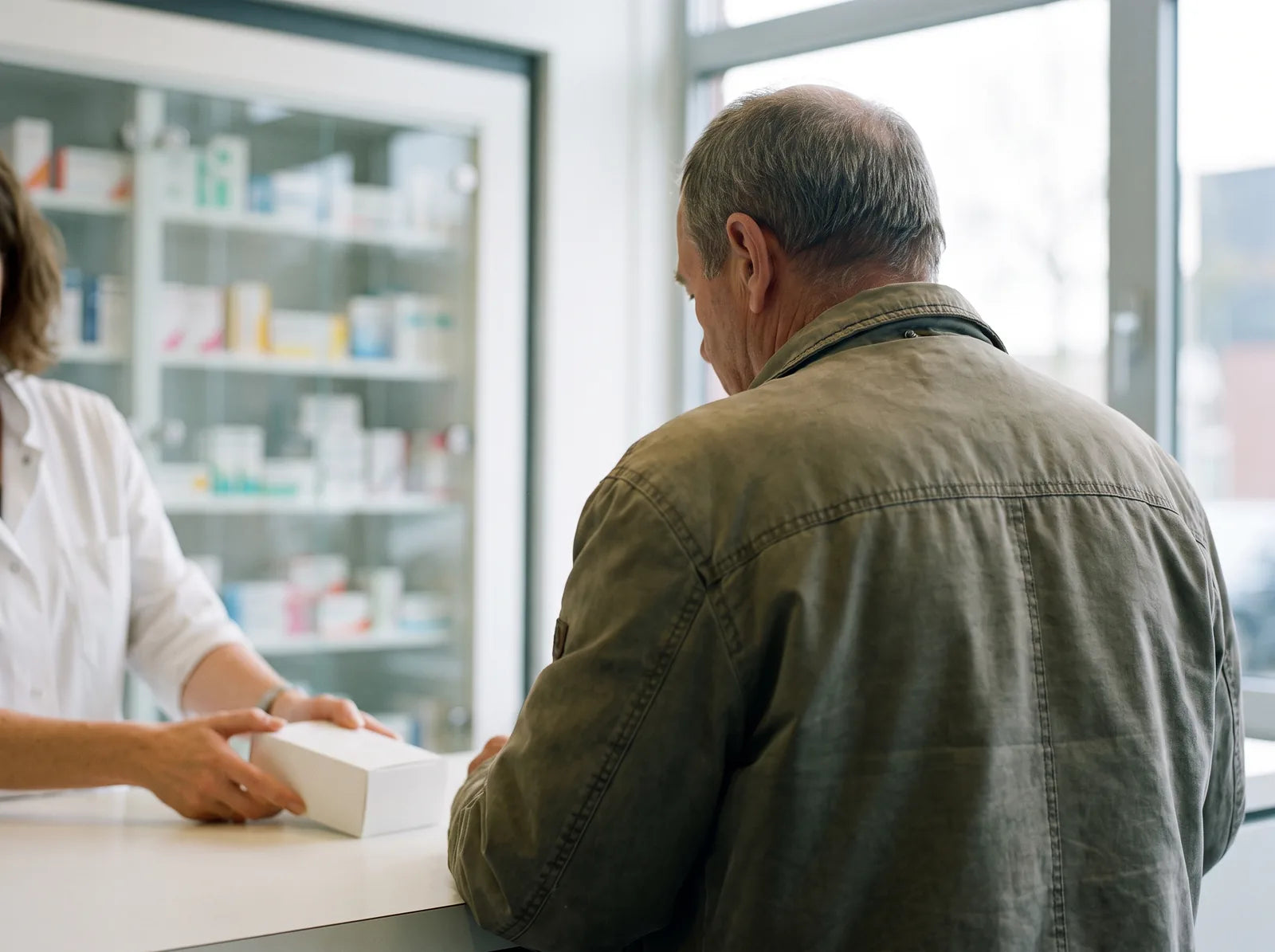 Sfeerbeeld: een man van achteren aan de apotheekbalie, de apotheker schuift een doosje toe