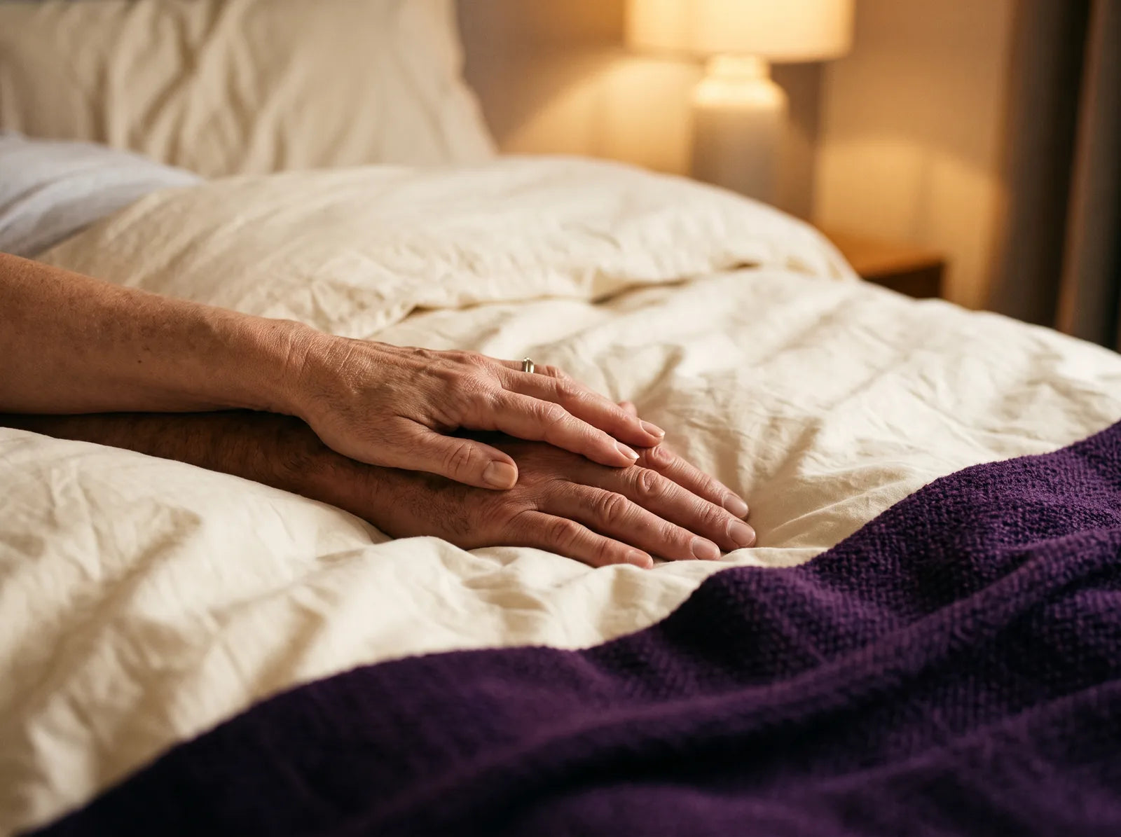 Sfeerbeeld: haar hand rust op de zijne op een crèmekleurig dekbed bij lamplicht