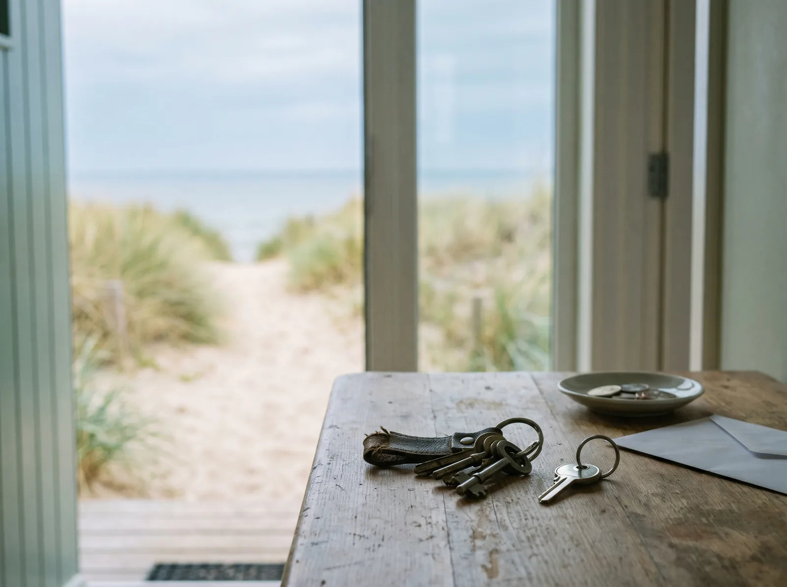 Sfeerbeeld: een oude sleutelbos en een nieuwe sleutel op een houten tafel bij een open deur naar de duinen