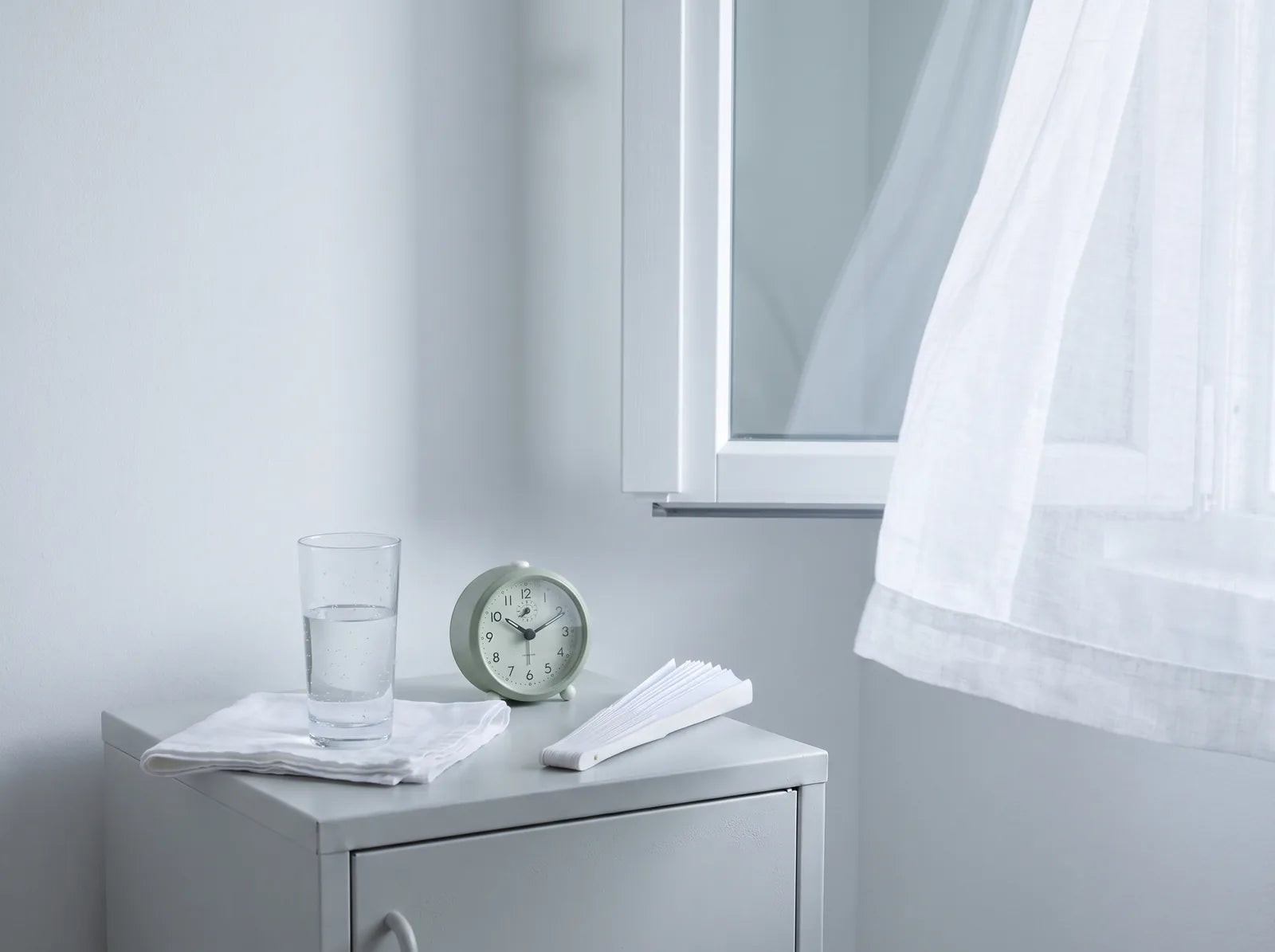 Sfeerbeeld: een nachtkastje met een glas water, een wekker en een waaier bij een open raam