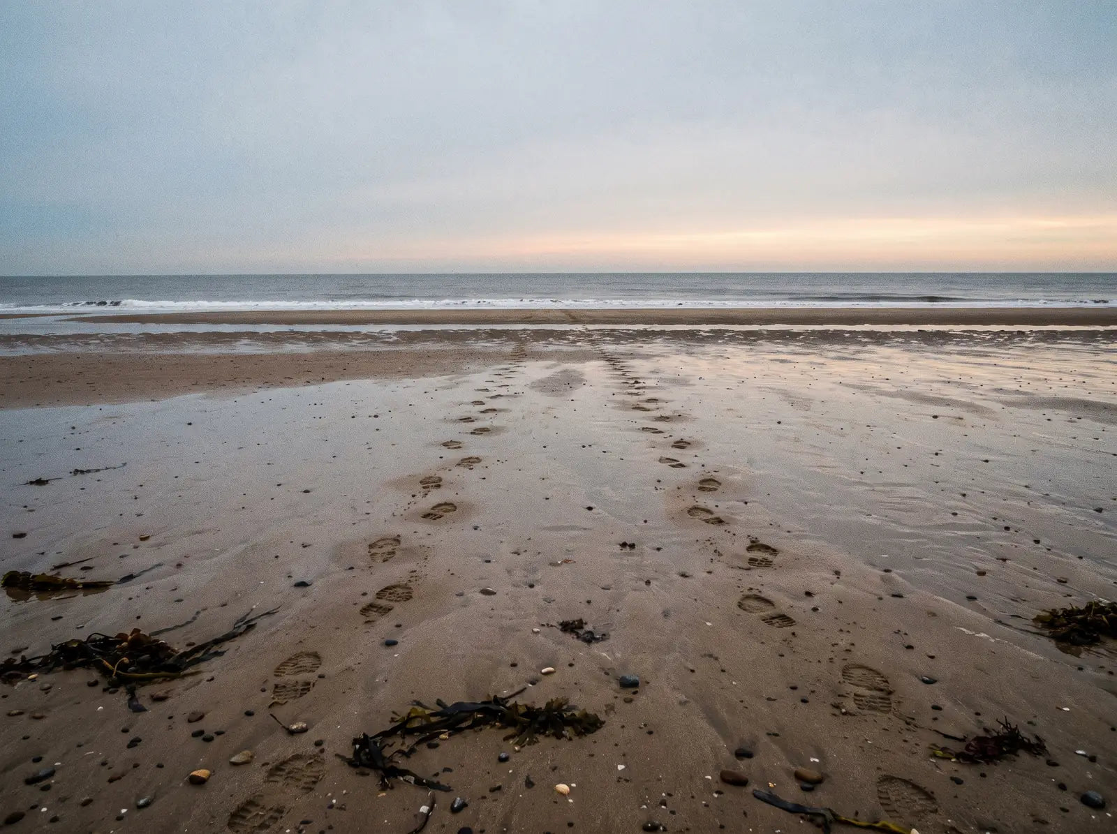 Sfeerbeeld: twee rijen voetstappen naast elkaar in het natte zand van een leeg strand bij eb