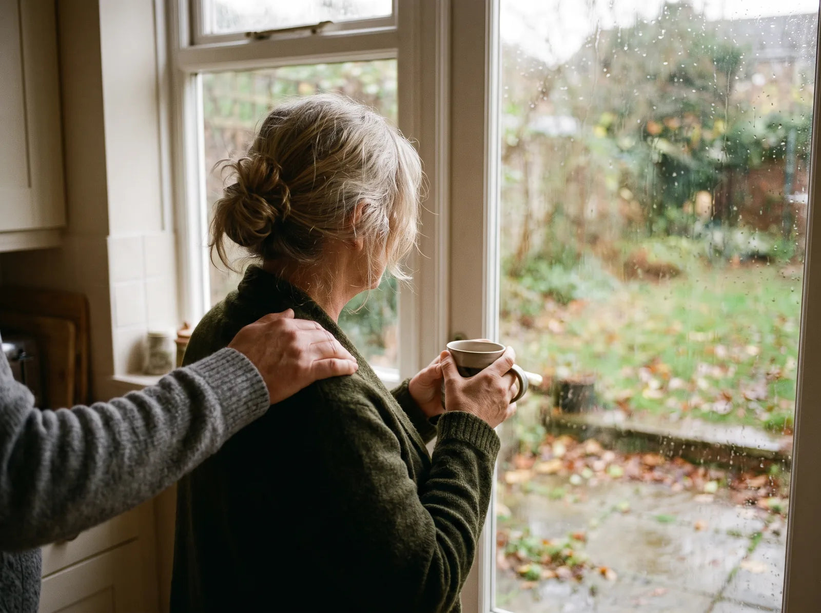 Sfeerbeeld: een vrouw van achteren bij het keukenraam met een kop thee, een hand op haar schouder