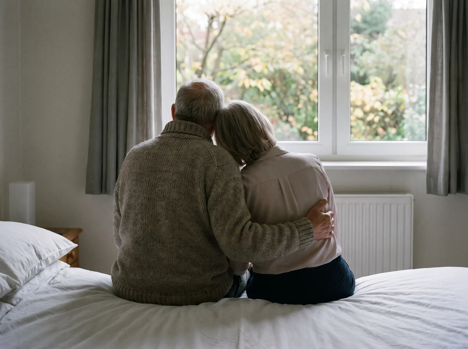 Sfeerbeeld: een stel van achteren op de rand van het bed, kijkend naar de herfsttuin
