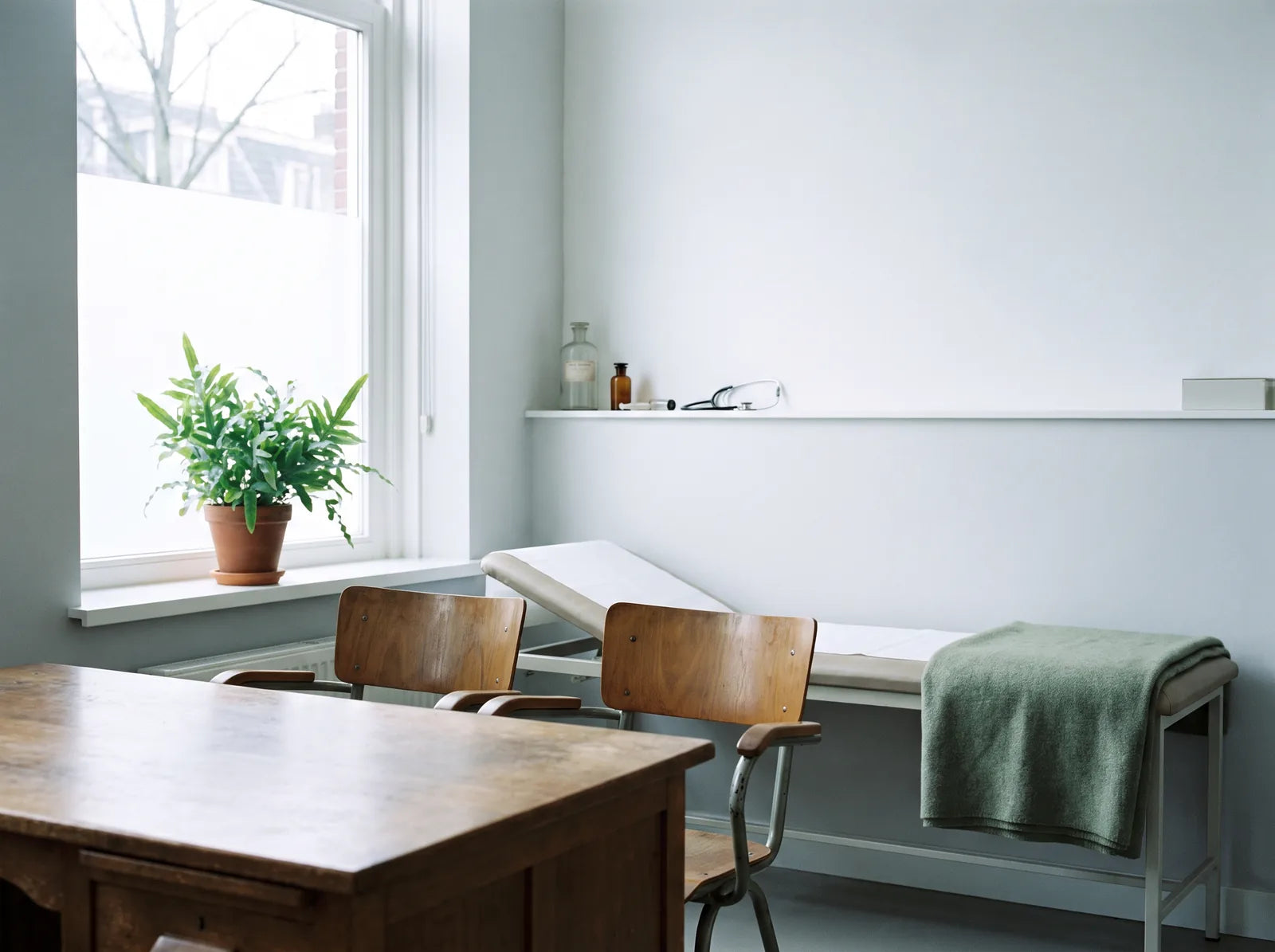 Sfeerbeeld: een lichte spreekkamer van een huisarts met een bureau, twee stoelen en een behandelbank