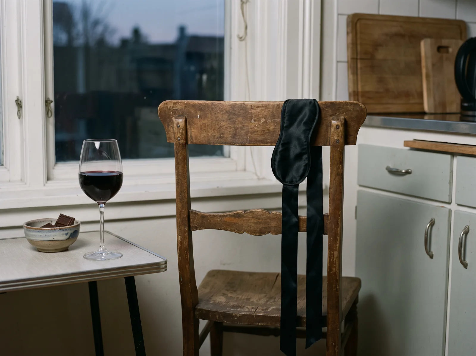 Sfeerbeeld: een zwarte satijnen blinddoek over een houten keukenstoel, met rode wijn en chocolade