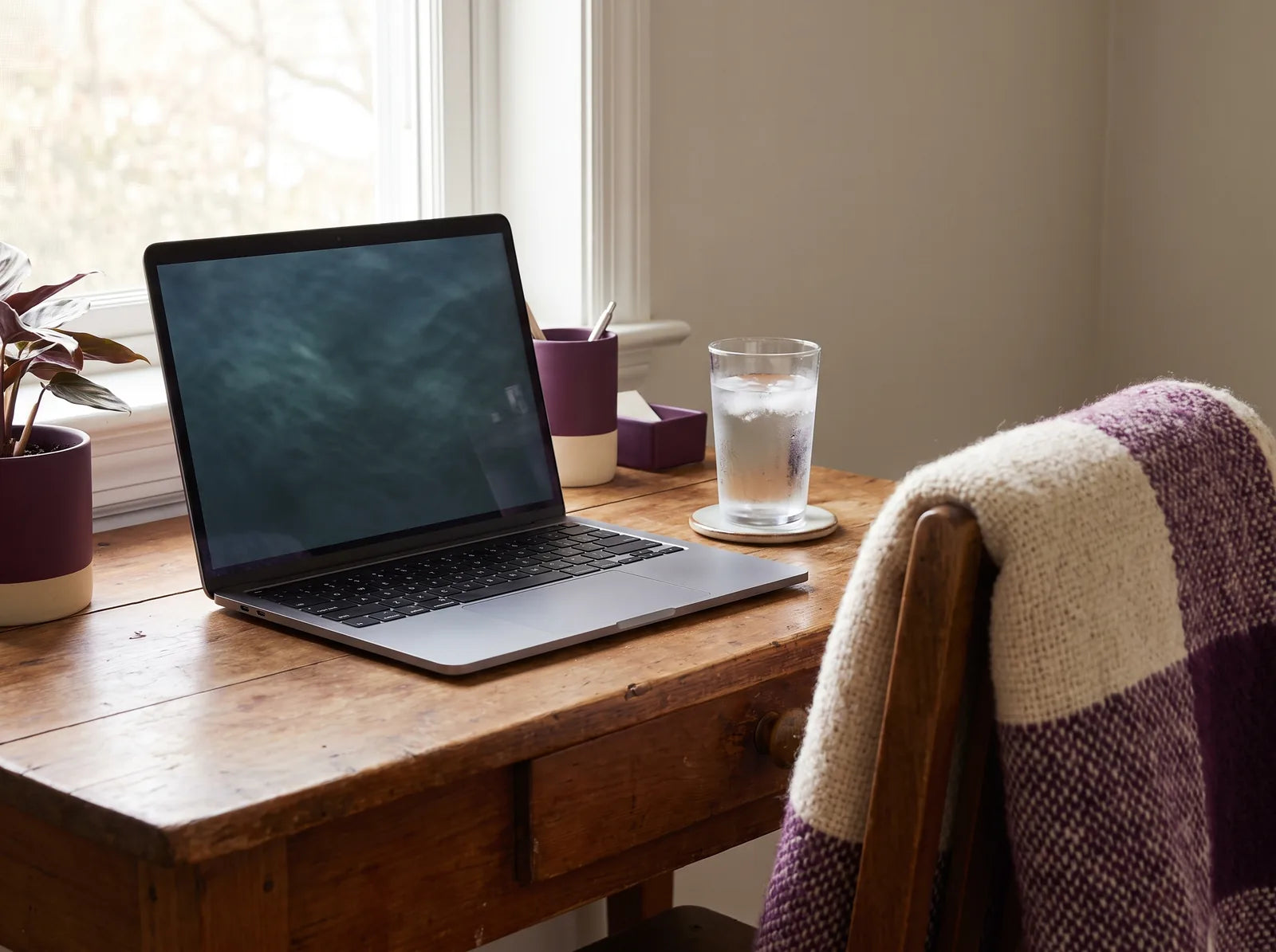 Sfeerbeeld: een laptop op een houten bureau bij het raam, met een glas water en een paarse plaid over de stoel