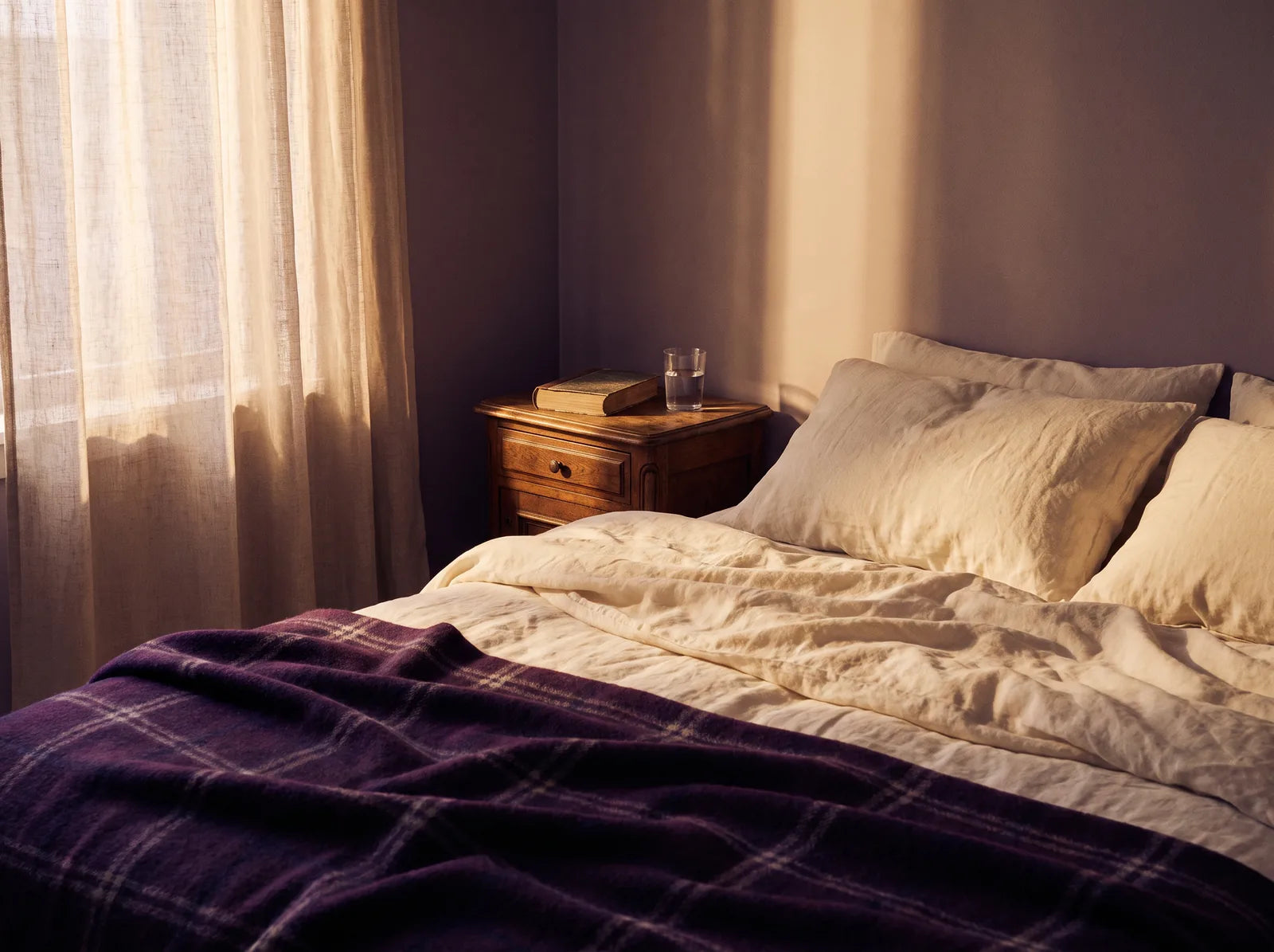 Sfeerbeeld: een rustige slaapkamer in ochtendlicht met creme beddengoed en een paarse plaid