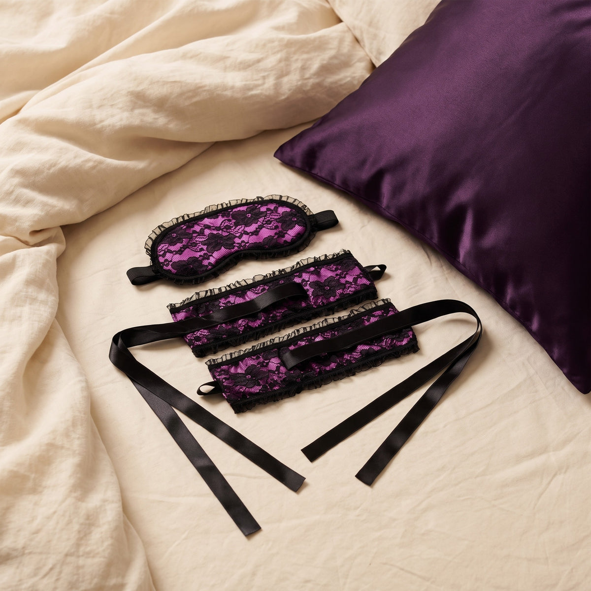 Sfeerbeeld: Burlesque blinddoek en polsboeien van satijn en kant op creme linnen naast een paars kussen