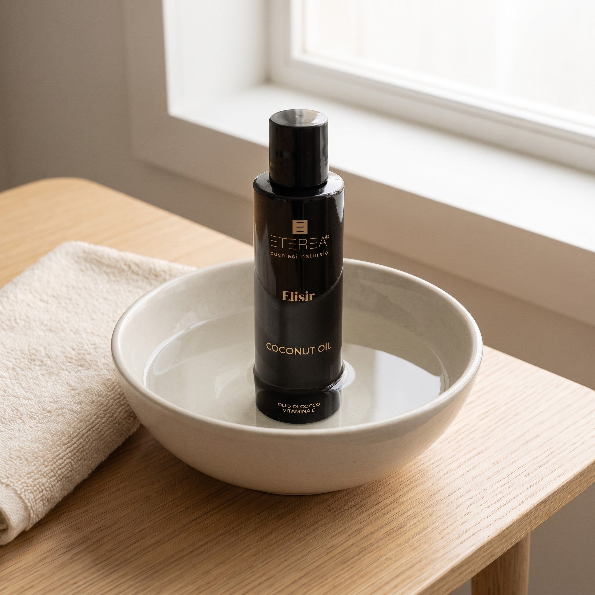 Fles Eterea Coconut Oil rechtop in een kom warm water op een houten tafel, met een handdoek ernaast