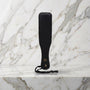 Kleine zwarte spanking paddle met hanglus rechtop op een marmeren plank, Fifty Shades