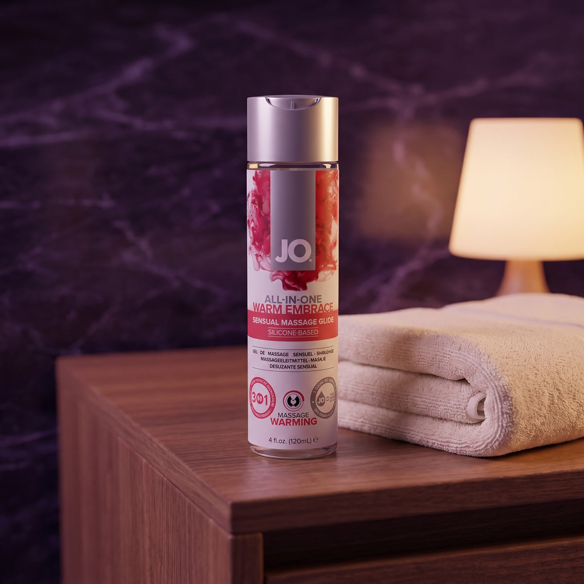 Sfeerbeeld: fles JO All-in-One Warm Embrace op een houten kastje met handdoek en lamp