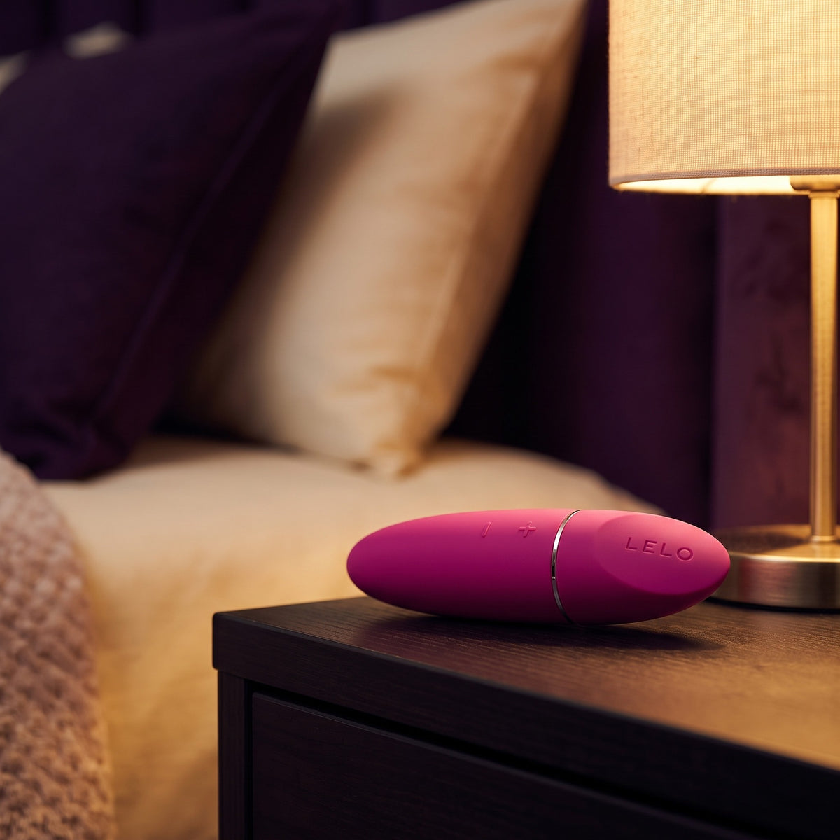 Sfeerbeeld: fuchsia roze Lelo Mia 3 liggend op een houten nachtkastje bij een brandende lamp