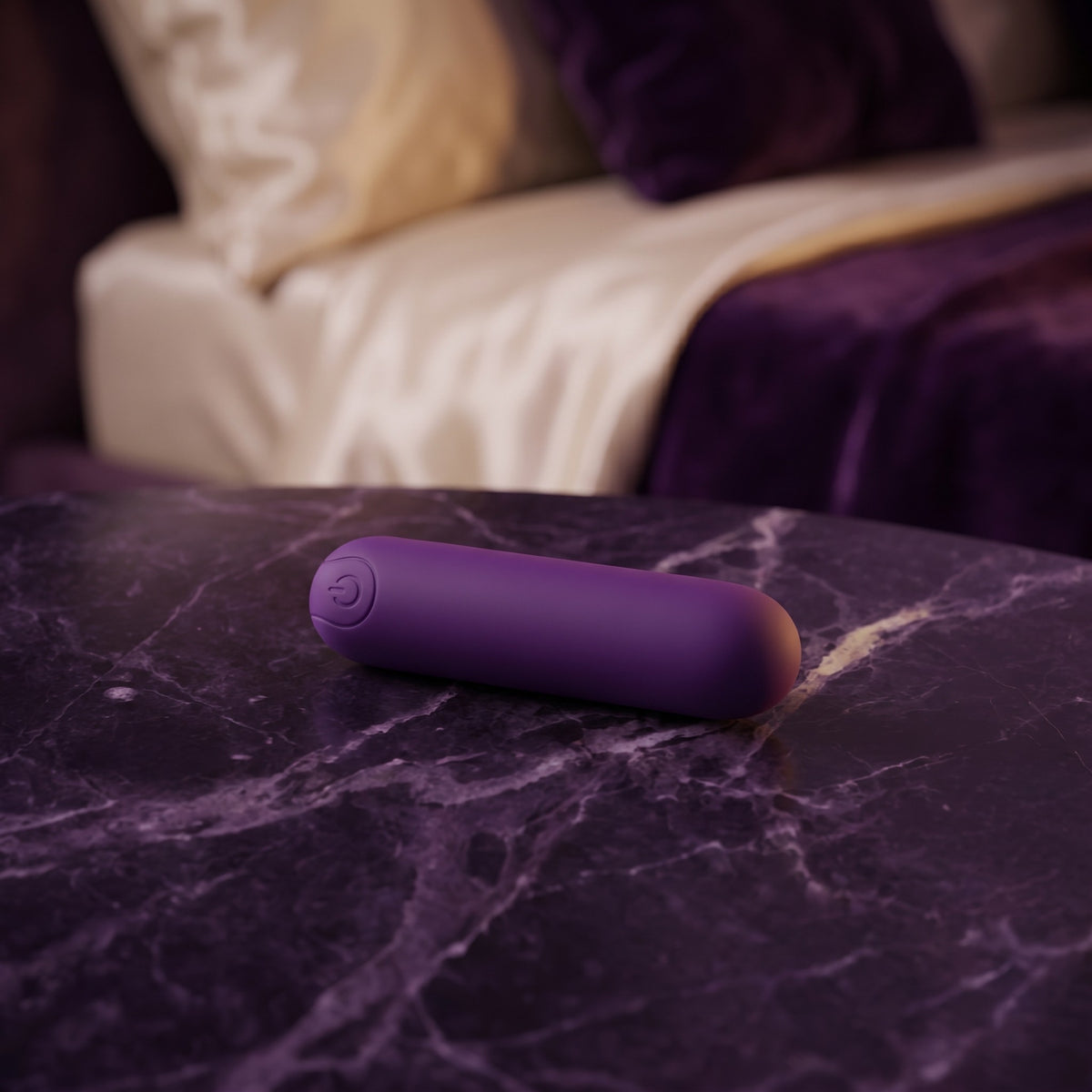 Sfeerbeeld: paarse bullet vibrator op donkerpaars marmer naast een bed in gedempt licht