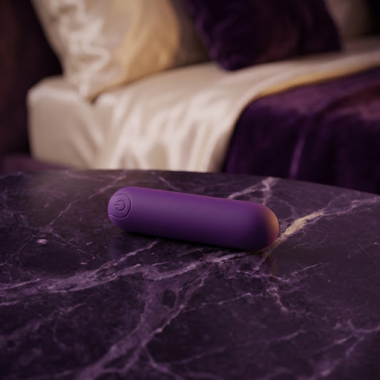 Sfeerbeeld: paarse bullet vibrator op donkerpaars marmer naast een bed in gedempt licht