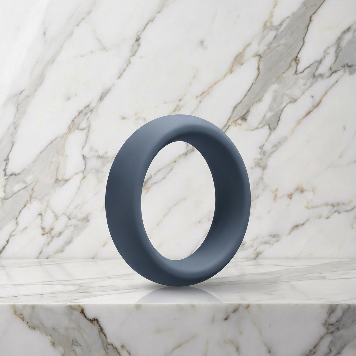 Brede leigrijze siliconen cockring van 45 mm op een marmeren plank, OptiMALE C-Ring