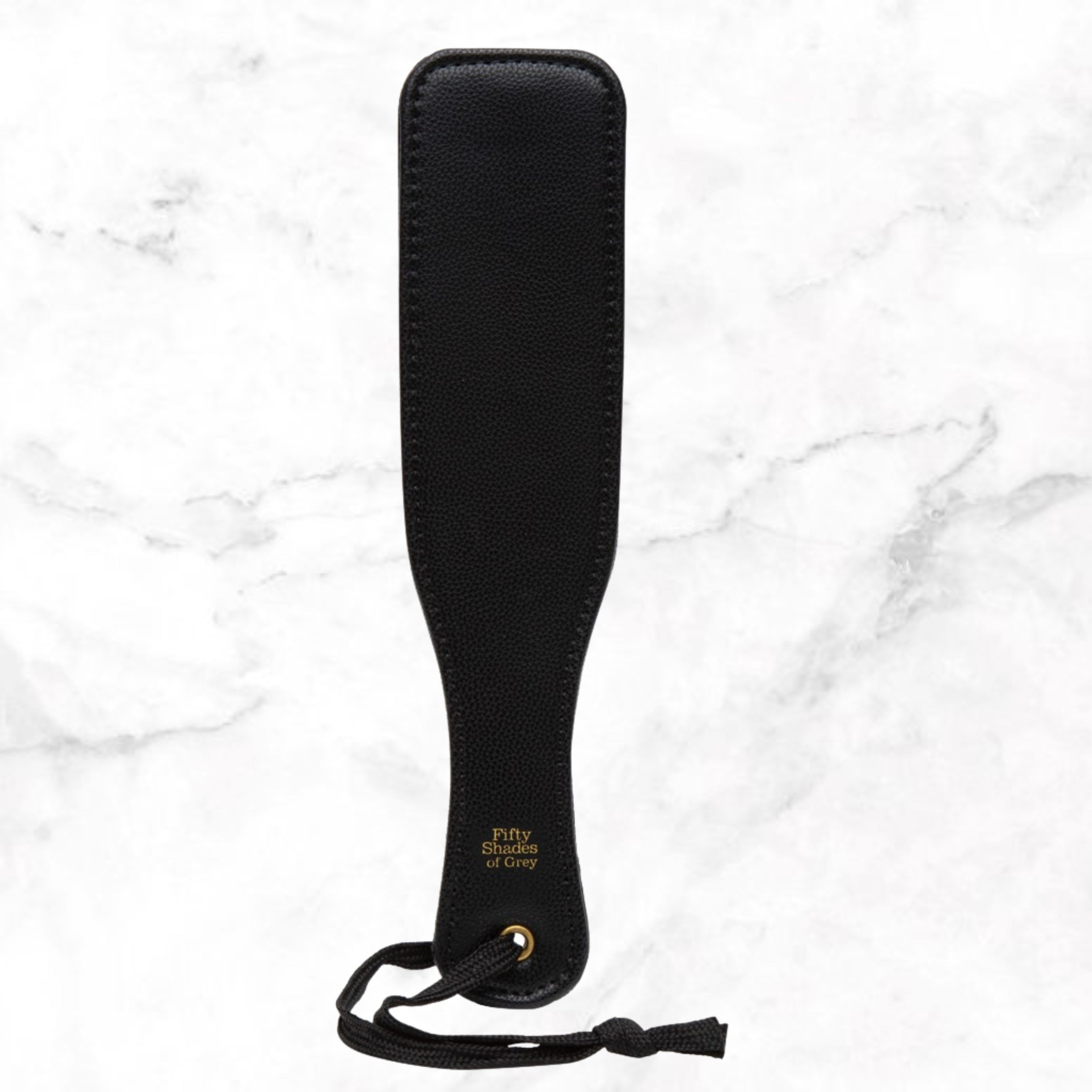 Zwarte paddle met goudkleurig logo en koordje op wit marmer, Fifty Shades of Grey
