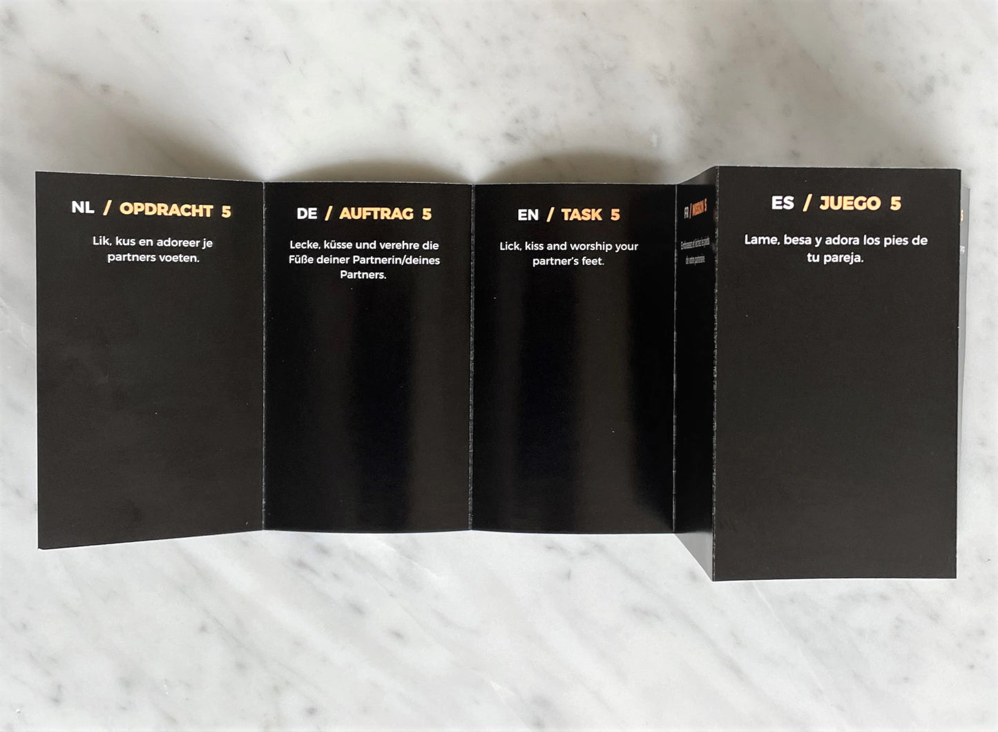 Uitgevouwen zwarte PleaseMe opdrachtkaart met opdracht 5 in vier talen op marmeren ondergrond