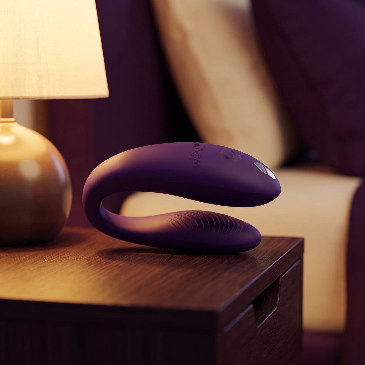 Sfeerbeeld: paarse We-Vibe Sync Go op een houten nachtkastje naast een warme lamp